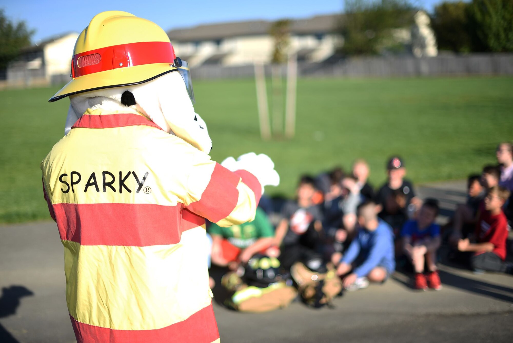 Sparky the Fire Dog, the mascot, communicates with grade school-aged children who are sitting. It is a sunny morning.