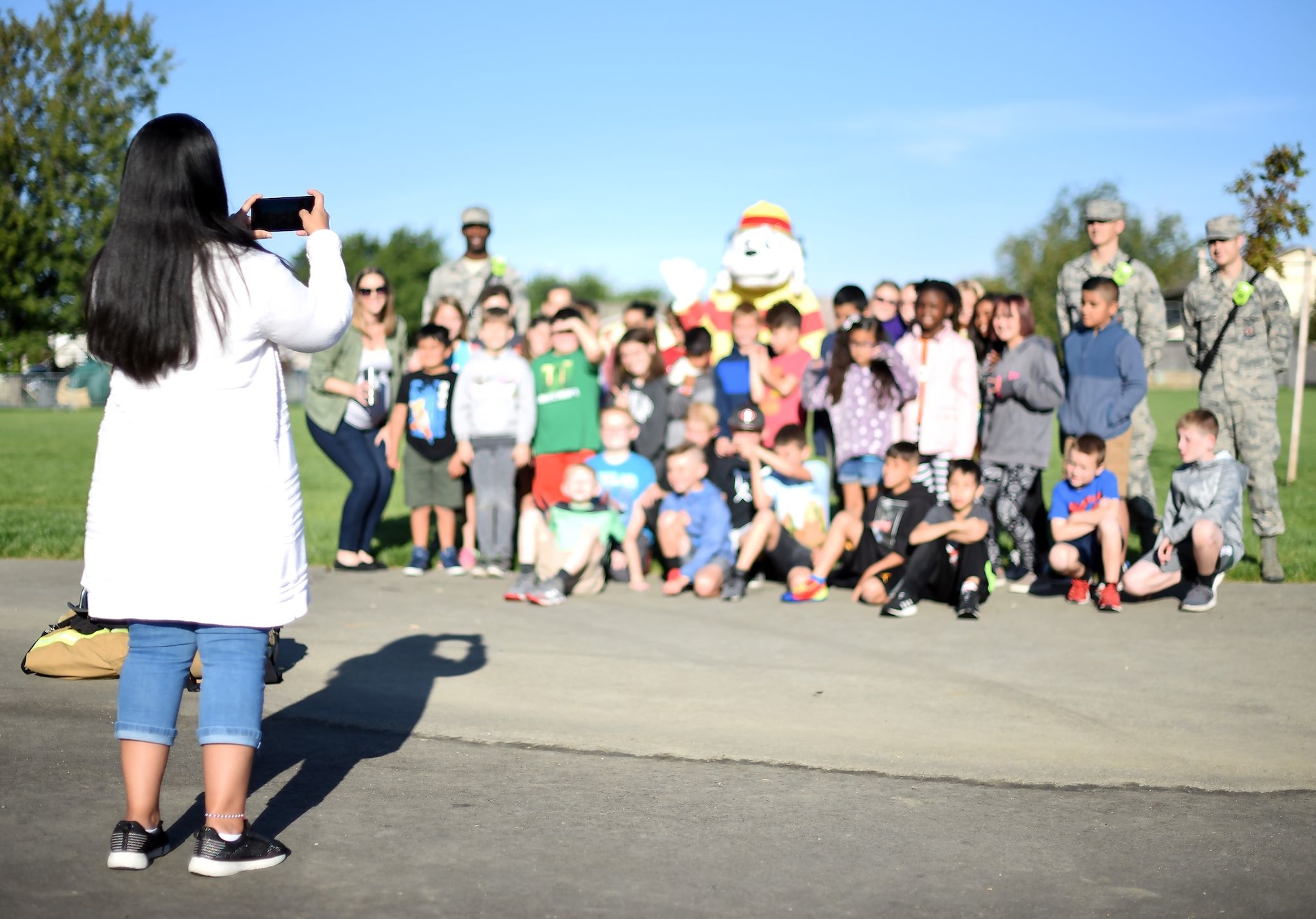 Grade school-aged children sit and stand among uniformed members of the Air Force along with Sparky the Fire Dog, a mascot for Fire Prevention Week, and pose for a photo taken by a smartphone.