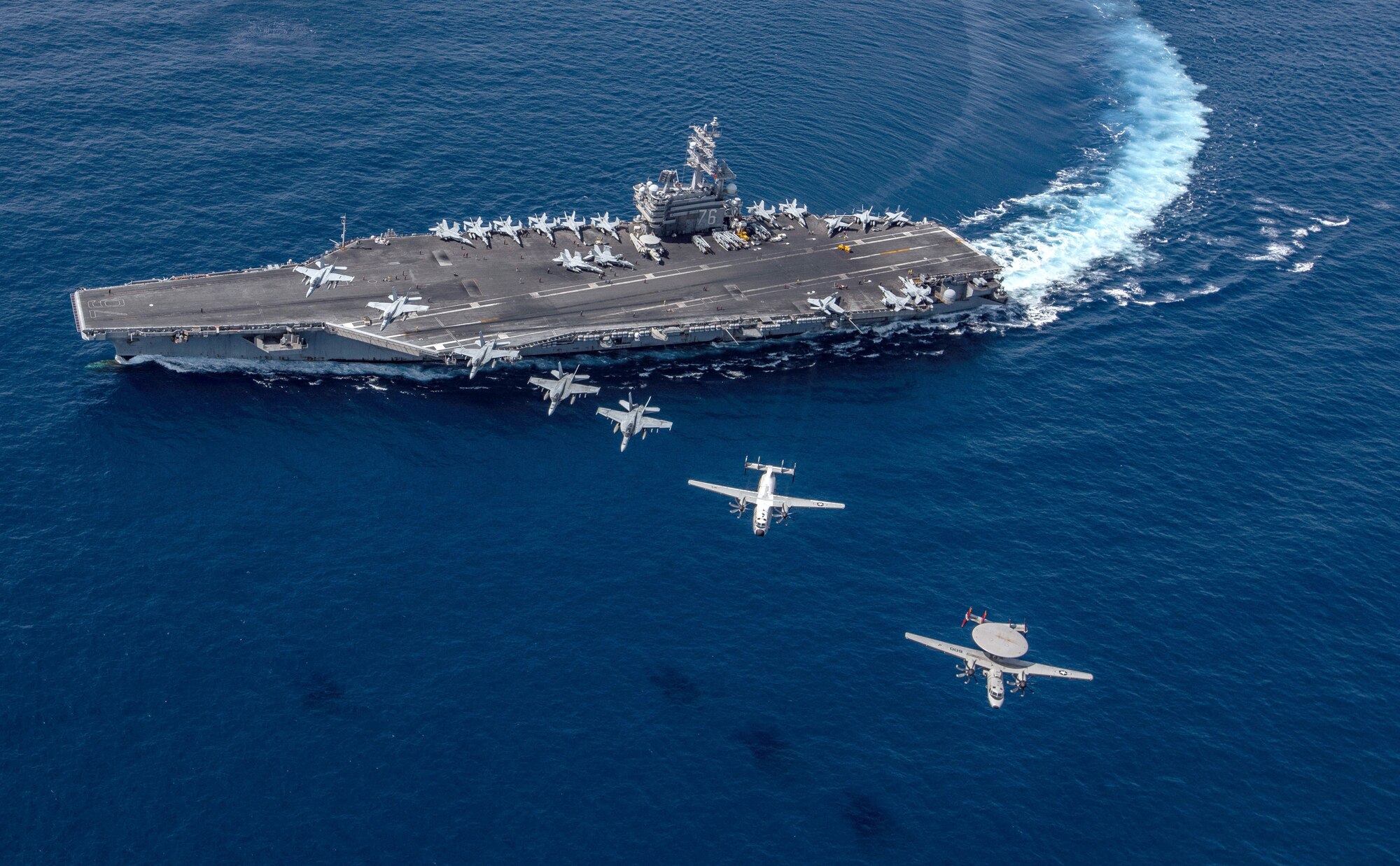 A group of aircraft fly in a line over a sailing aircraft carrier.