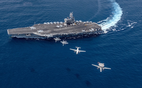 A group of aircraft fly in a line over a sailing aircraft carrier.