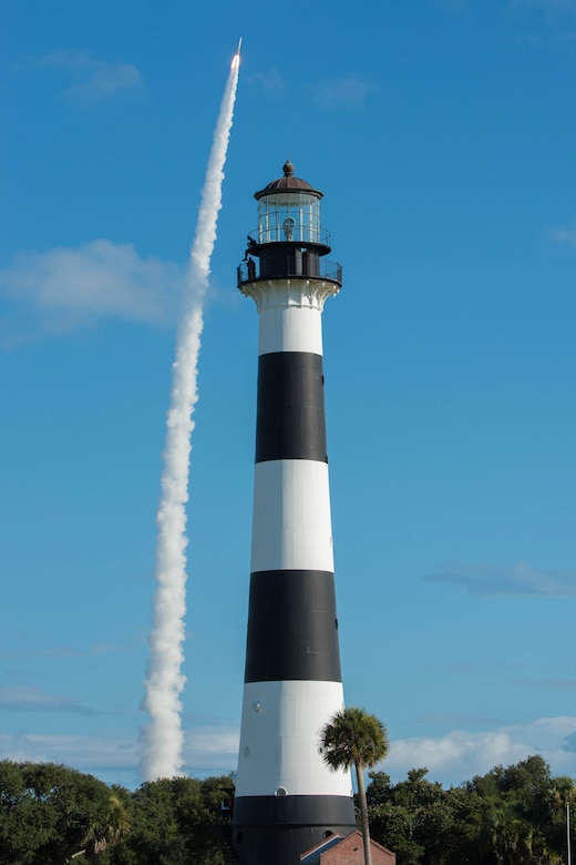 A rocket launches into the sky behind a tall black and white lighthouse.