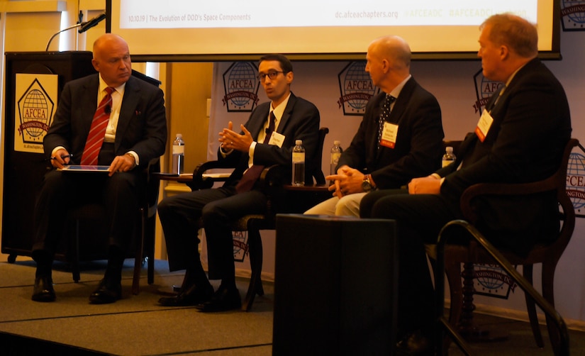 Man seated among three others on a panel gestures while speaking.