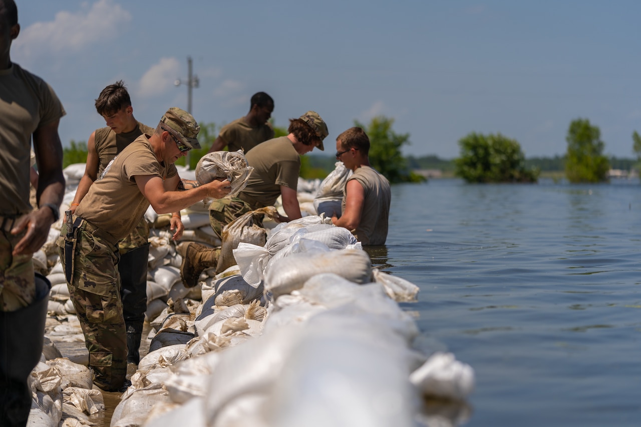 A guardsman standing in waist-high water works with other service members to place sandbags on a levee.