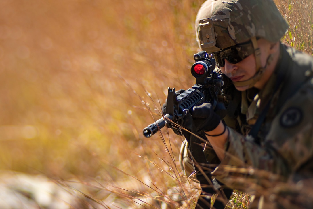 A soldier crouched in field of tall grass aims a weapon.