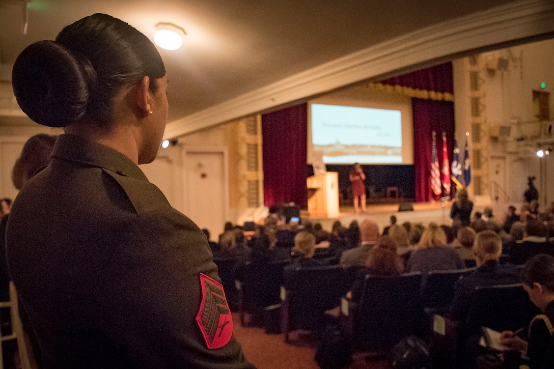 A Marine stands near the rear of an auditorium as a speaker addresses an audience.