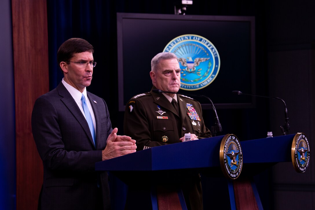 Two men, one in a suit, and one in a military uniform, stand behind lecterns. The lecterns bear the logo of the U.S. Defense Department.