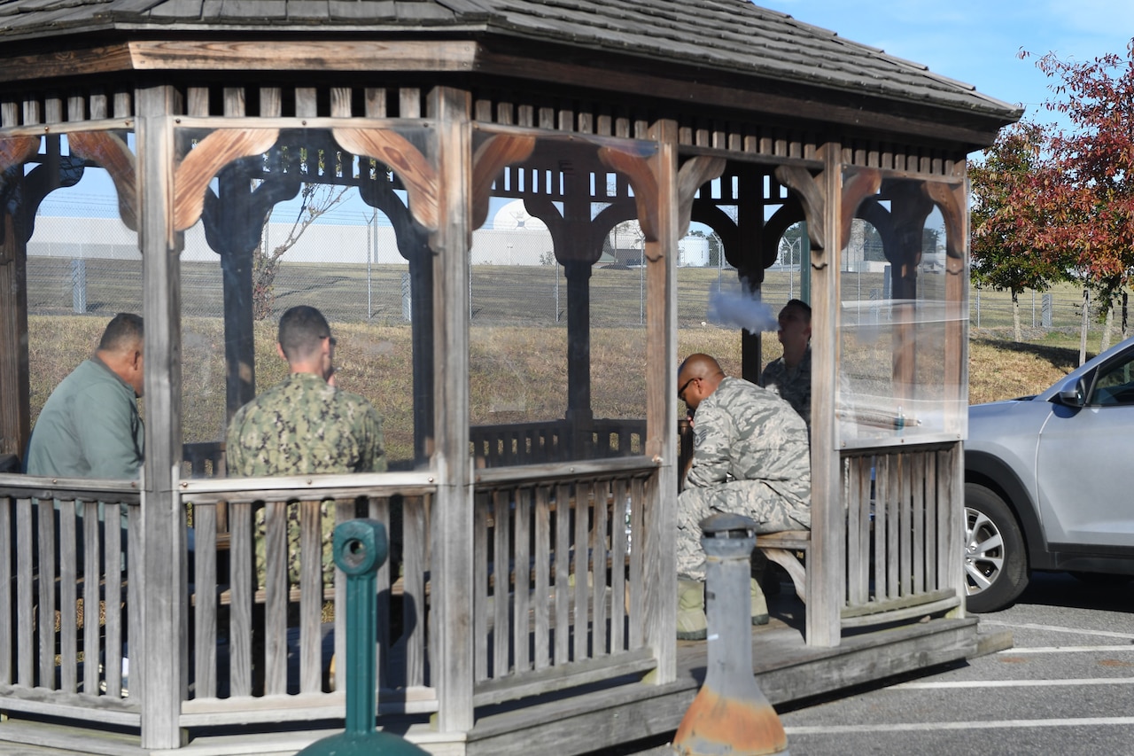 Military personnel sit under a wooden structure while using tobacco or vape products.