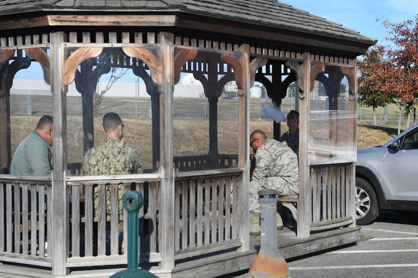 Military personnel sit under a wooden structure while using tobacco or vape products.