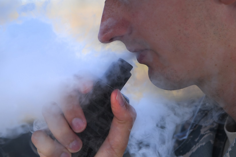 A service member holds a vape pipe near his mouth. A cloud of vapor surrounds his face.