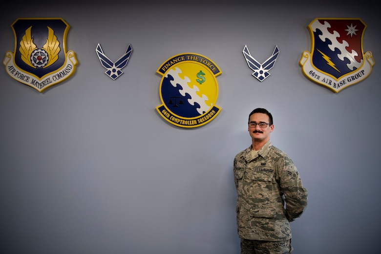 Airman 1st Class Aws Hussein, 66th Comptroller Squadron financial management technician, stands under the Air Force Material Command, 66th Air Base Group and CPTS crests on Hanscom Air Force Base, Massachusetts, Oct. 10. Originally from Baghdad, Iraq, Hussein worked alongside coalition forces as an Arabic interpreter from 2007 to 2011. He immigrated to the U.S. with his wife and two young children before enlisting in the Air Force earlier this year.