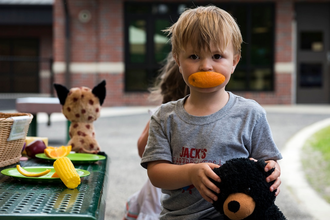 A boy holds a teddy bear and eats an orange.