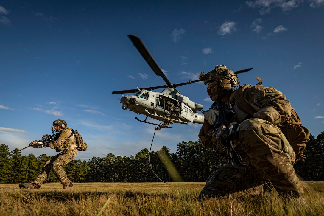 Two airmen maneuver in a field as a third hangs from a rope out of a hovering helicopter.
