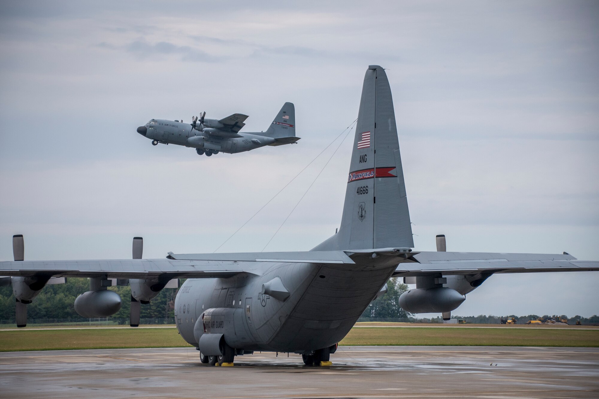 Photo of a C-130 flying over the flight line of another C-130