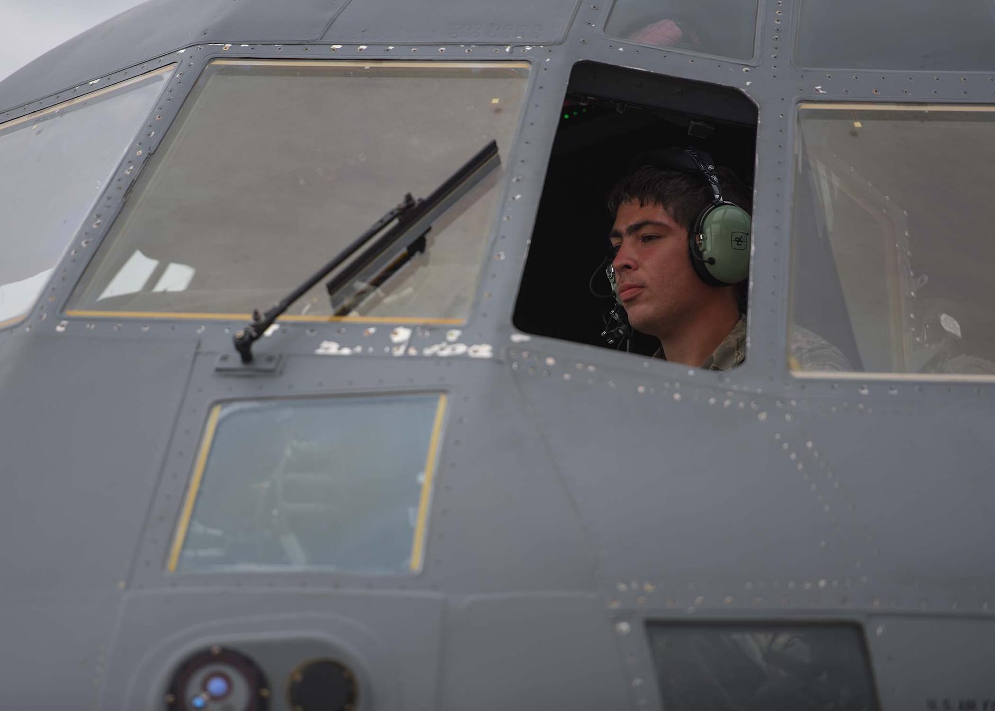 An Airman is seen through the window of a grey plane with a headset on.
