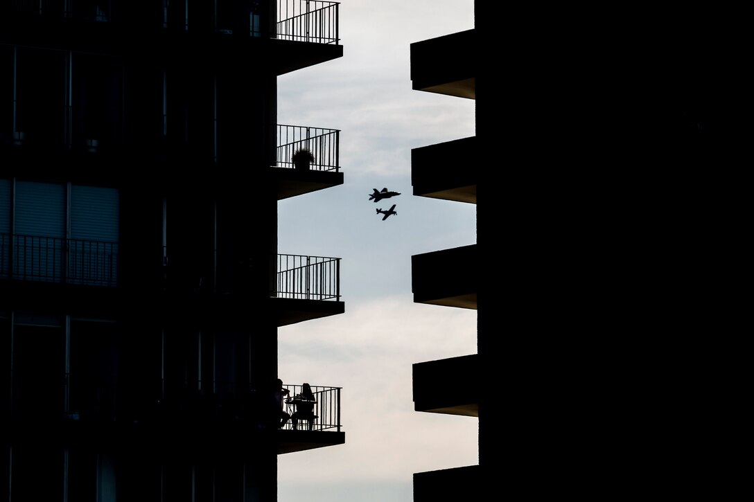 Silhouettes of two aircraft are visible through a zigzag-shaped space between two buildings with protruding balconies.