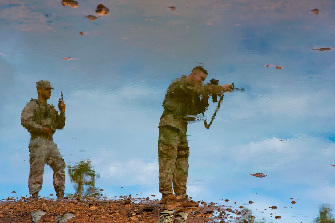 A view of two standing airmen, one pointing a weapon, is reflected in light blue water.