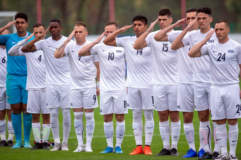 Military soccer players stand in a row and salute on a field.