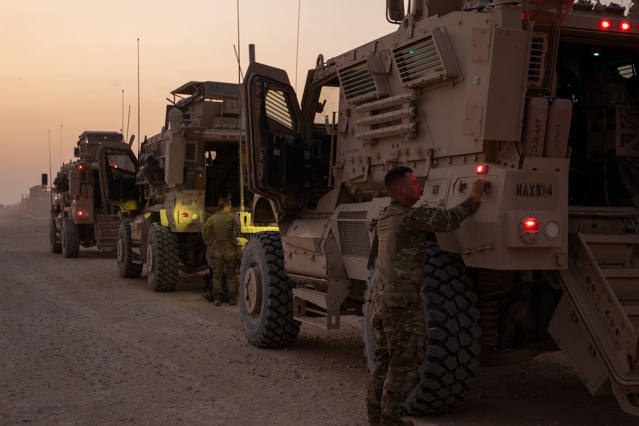 A soldier stands outside of a military vehicle; with other vehicles parked in front.