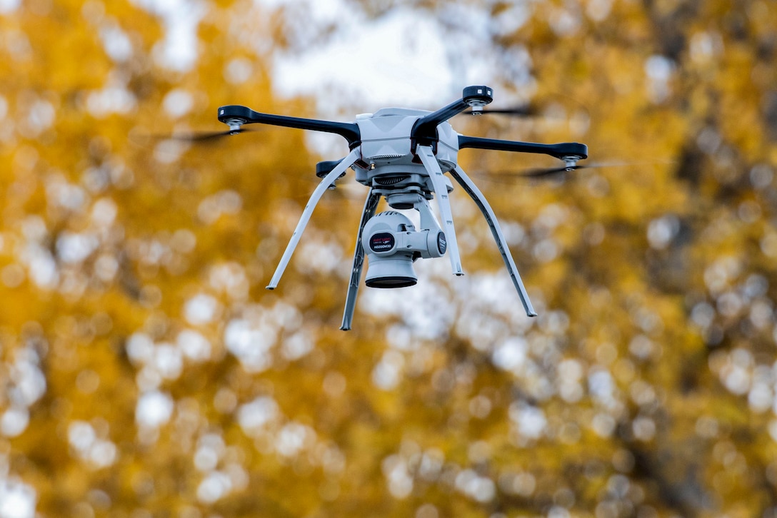 A drone hovers in the air, against a backdrop of trees with yellow leaves.