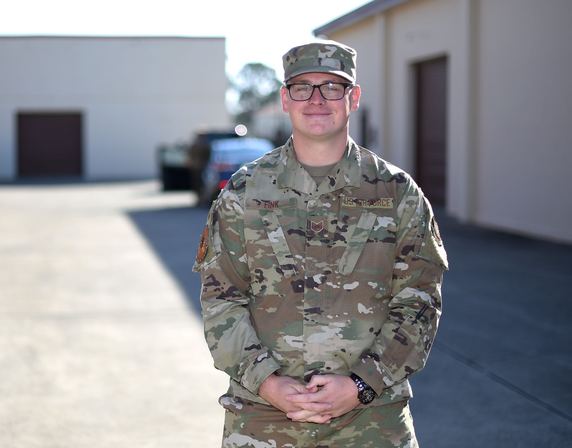 A uniformed Airman stand in a long. asphalt parking lot along an industrial-style warehouse on sunny day.