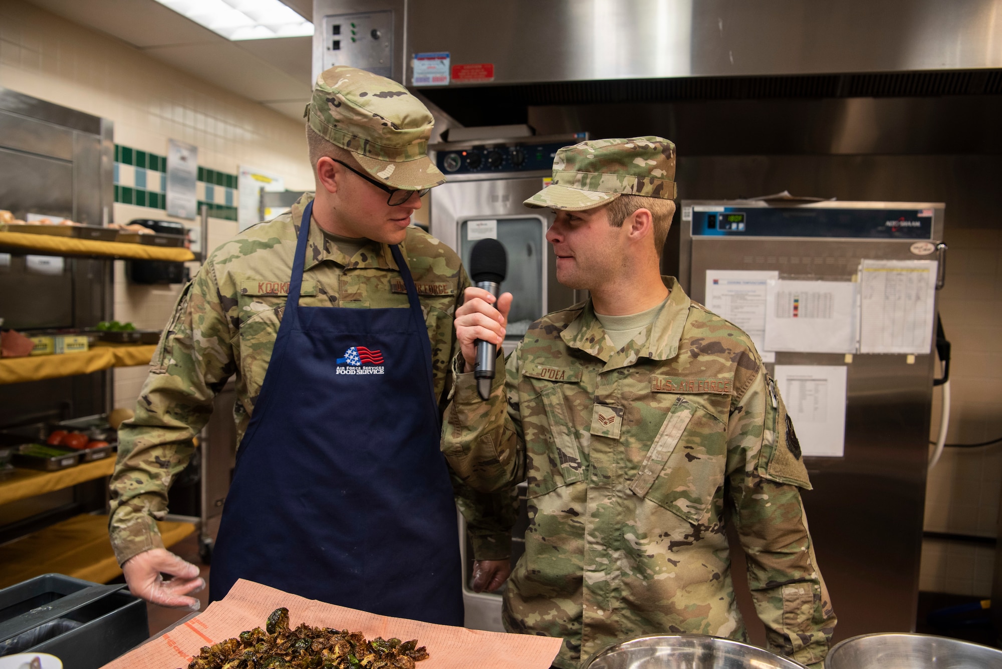 Senior Airman Kyle O’dea, 90th Force Support Squadron missile chef, interviews Airman 1st Class Nathaniel Kooker, 90th FSS food service specialist, about the items he has been preparing during a cooking competition Oct. 15, 2019, at F.E. Warren Air Force Base, Wyo. O’dea was one of the hosts during the competition and provided commentary for the living feed into the dining hall where the judges and fans sat. (U.S. Air Force photo by Senior Airman Abbigayle Williams)