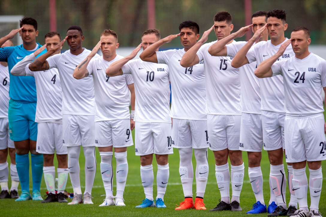 Military soccer players stand in a row and salute on a field.