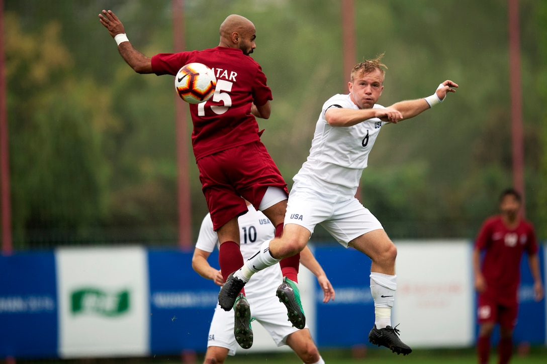 A U.S. Army soccer player and a Qatari soccer player leap in the air while facing each other on a field..