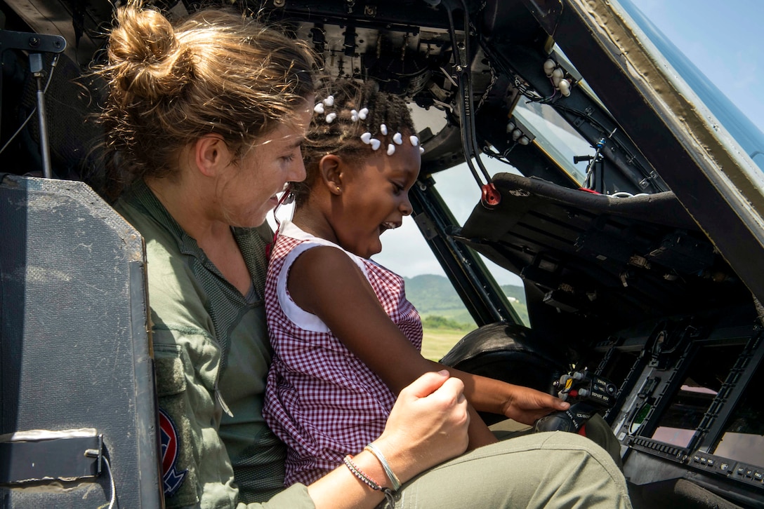 A girl sits on a sailor's lap and smiles as she plays with a helicopter's controls.