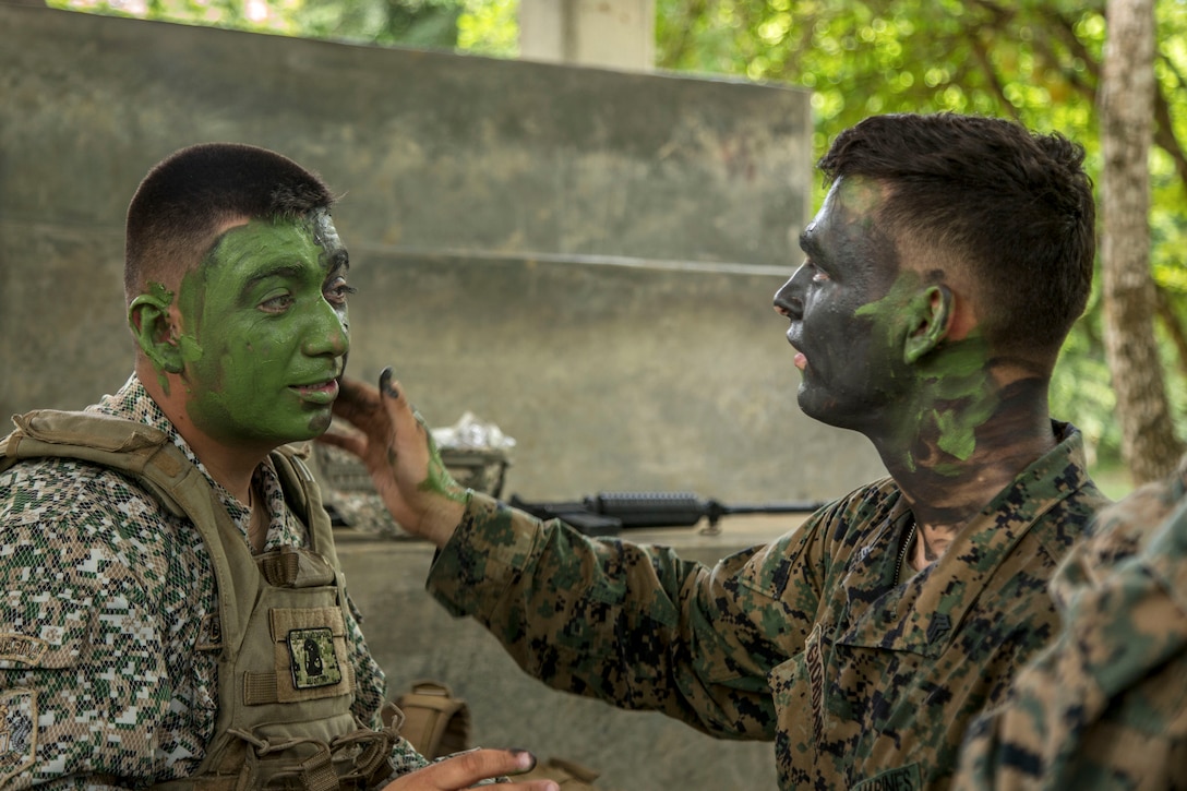 A Marine hand-applies camouflage face paint on a Colombian service member outside in a wooded area.