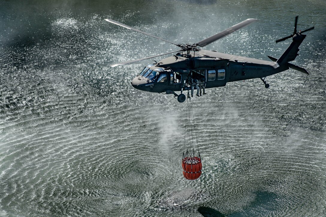A helicopter flies over a body of water with a bucket suspended underneath by a cable.