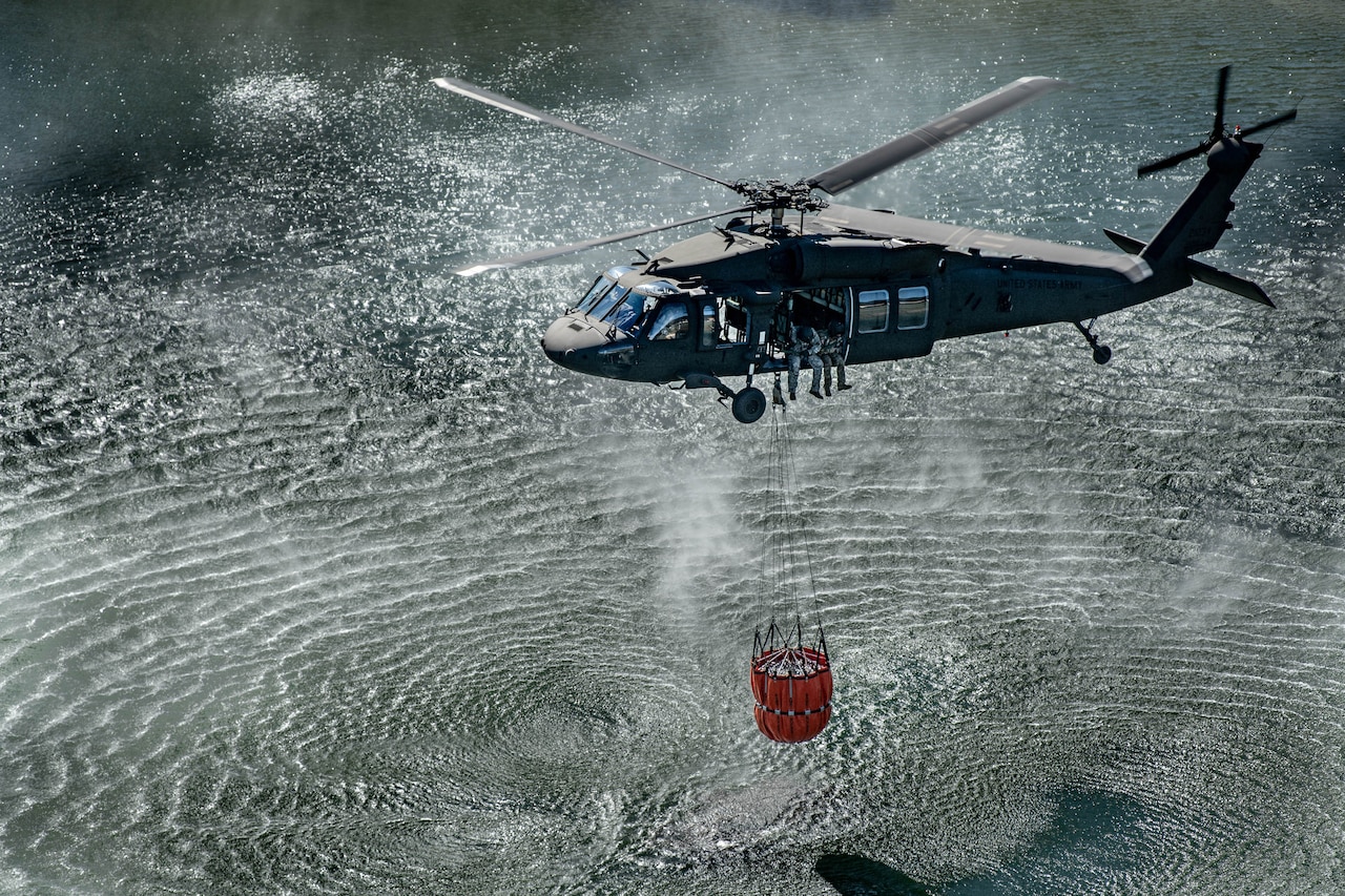 A helicopter flies over a body of water with a bucket suspended underneath by a cable.