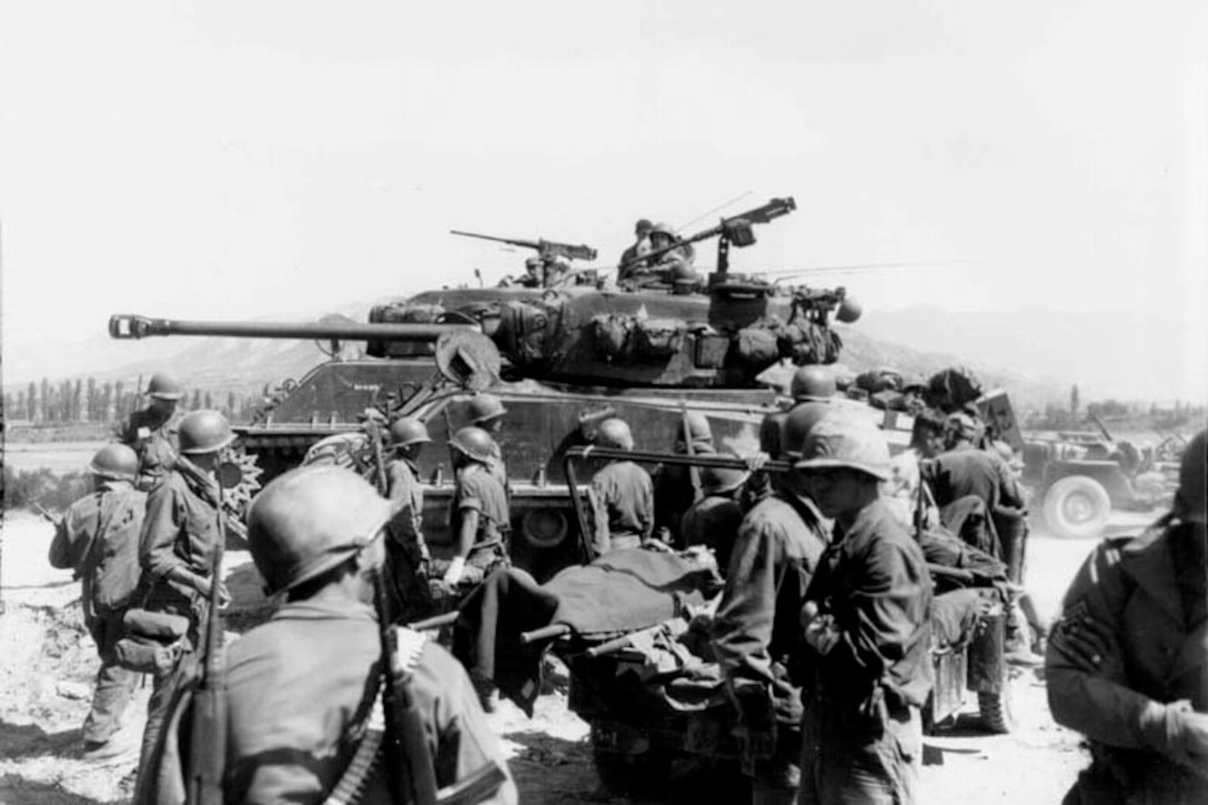 Several soldiers in protective helmets stand near an M-4 tank. One man is looking out from a hatch on top.