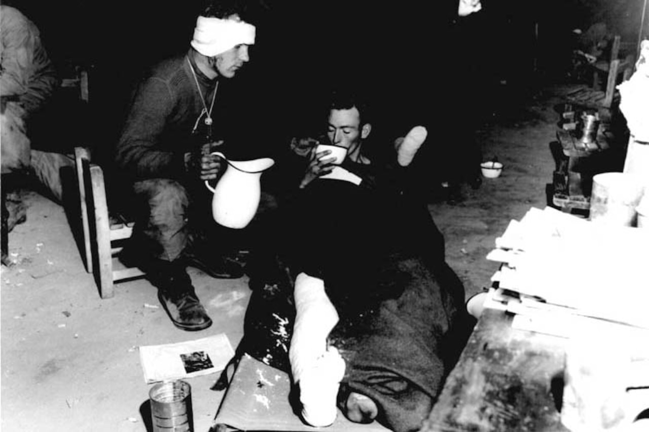 A wounded soldier, whose leg is bandaged, raises his head to sip from a large bowl while lying on the floor under a blanket. Another soldier, whose head is bandaged, sits beside him in a chair holding a pitcher and prepares to pour refill the bowl held by his wounded comrade.