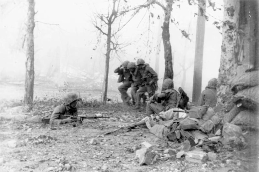 Two soldiers carry a third between them past sparse trees on a battlefield. In the foreground, three other soldiers holding weapons keep watch.