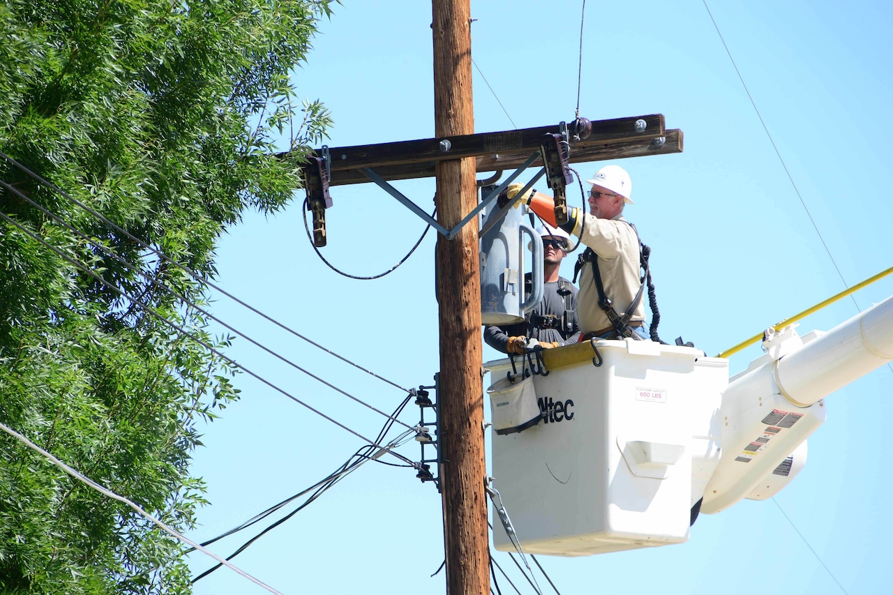Men in a bucket crane work on a power transformer that sits atop a wooden pole.