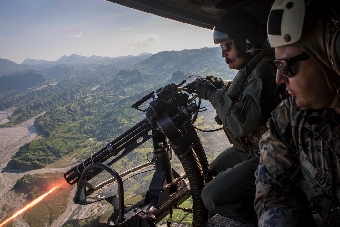 A Marine fires a weapon from a helicopter: another Marine sits beside.