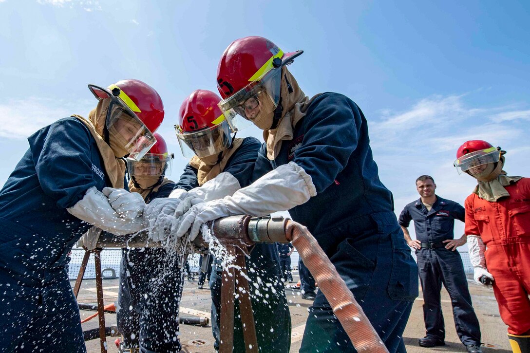 A group of sailors use their hands to stop water for pouring out of a hole in a water hose.