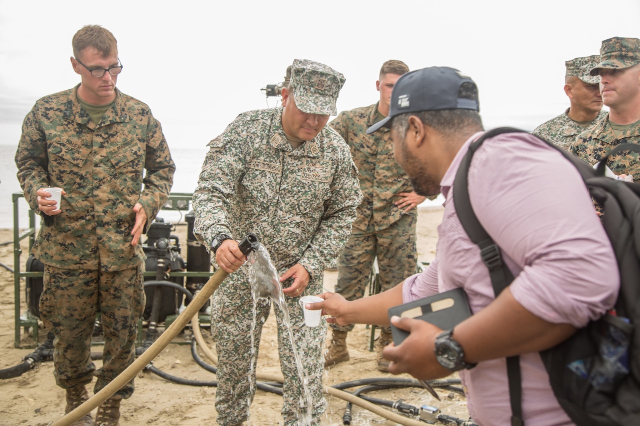 Colombian military officer fills a cup with water from a hose. A civilian is holding the cup, and Marines in uniform observe.