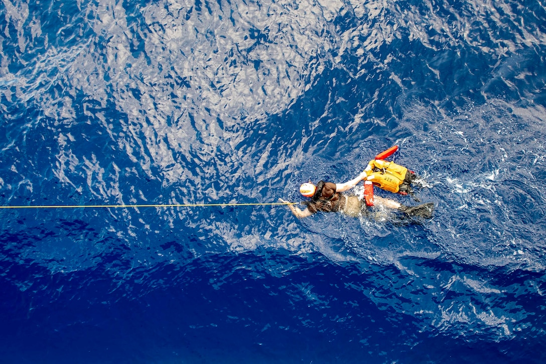 A sailor swims in bright blue water, holding a line and an inflated life vest.