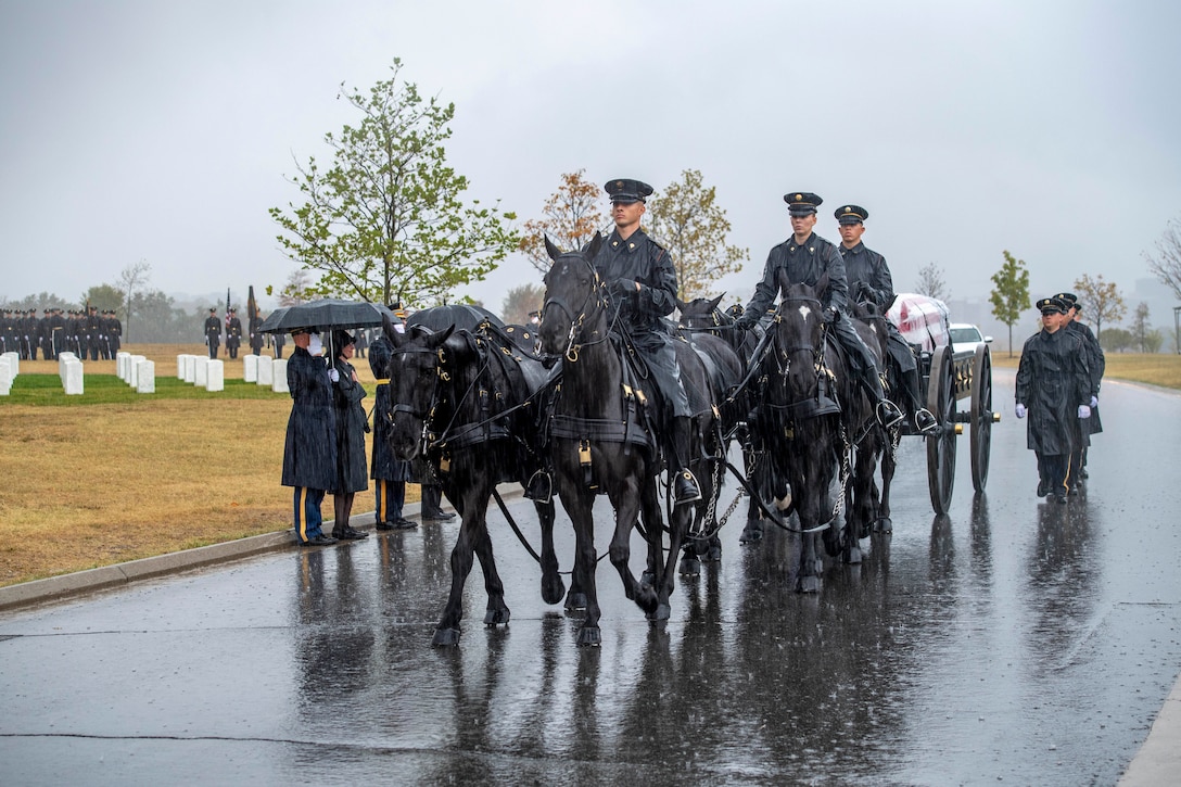 A group of soldiers and horses lead a funeral escort through a cemetery.