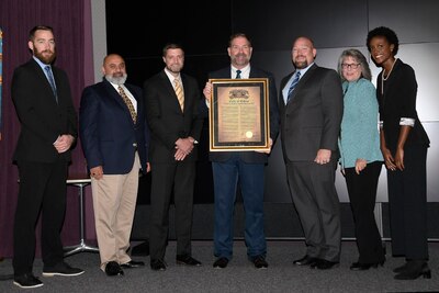Group of people stand for photo, middle person holds a frame