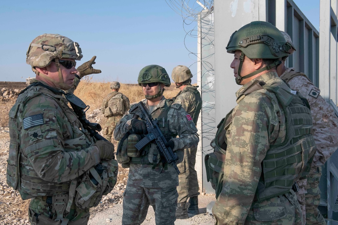 Soldiers in camouflage uniforms and protective gear talk near a wall.