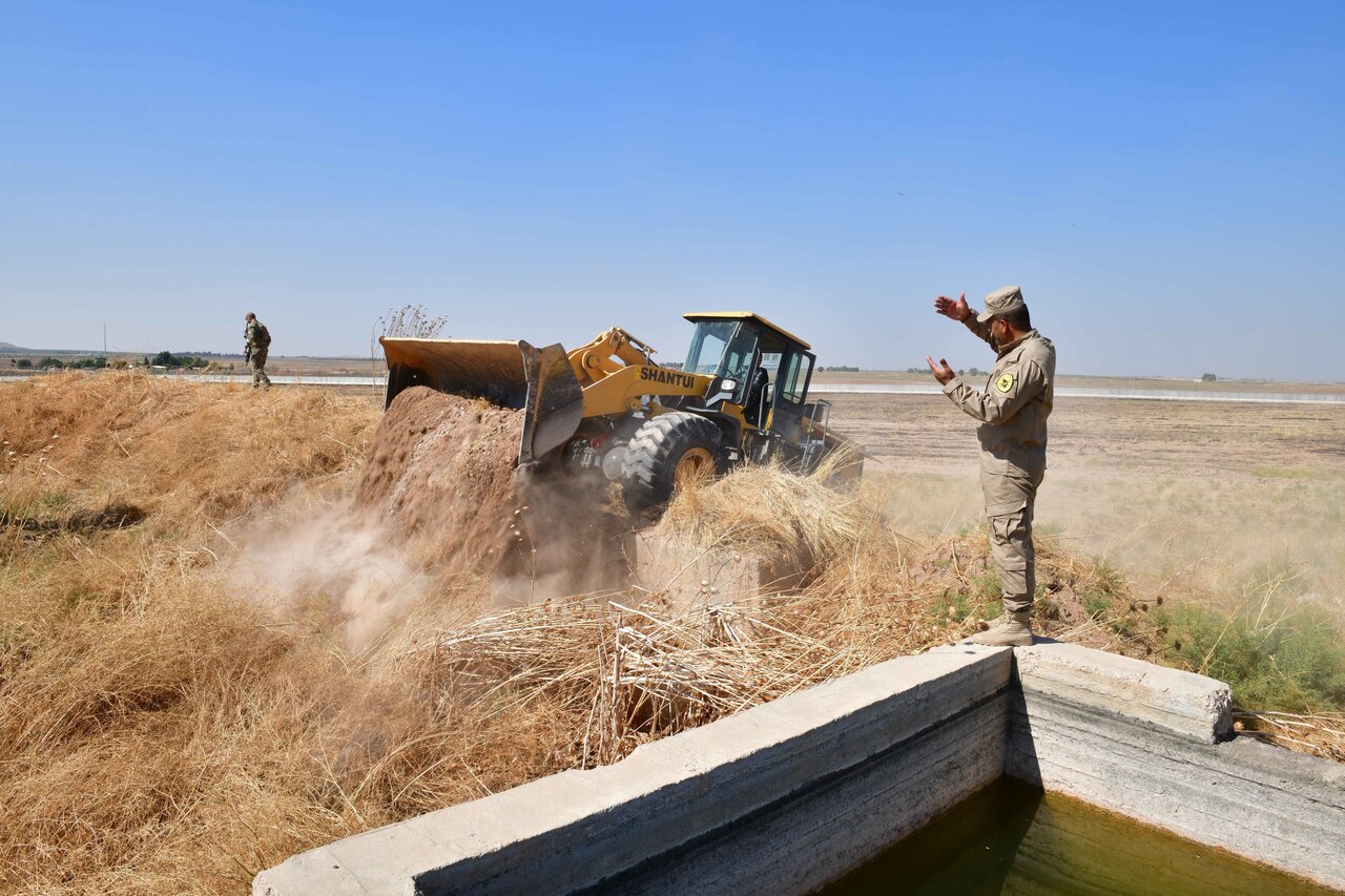A man in a camouflage uniform directs a backhoe driver dumping a load of dirt.