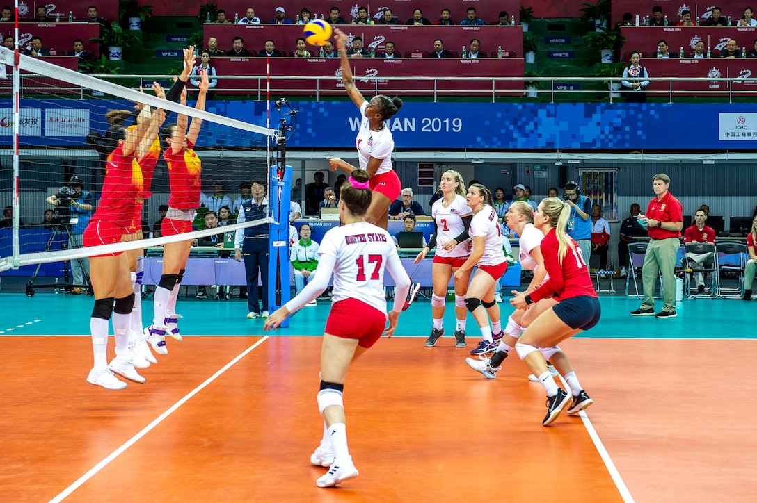 A U.S. athlete leaps and hits a ball as her teammates look on and opponents raise their arms on the other side of the net.