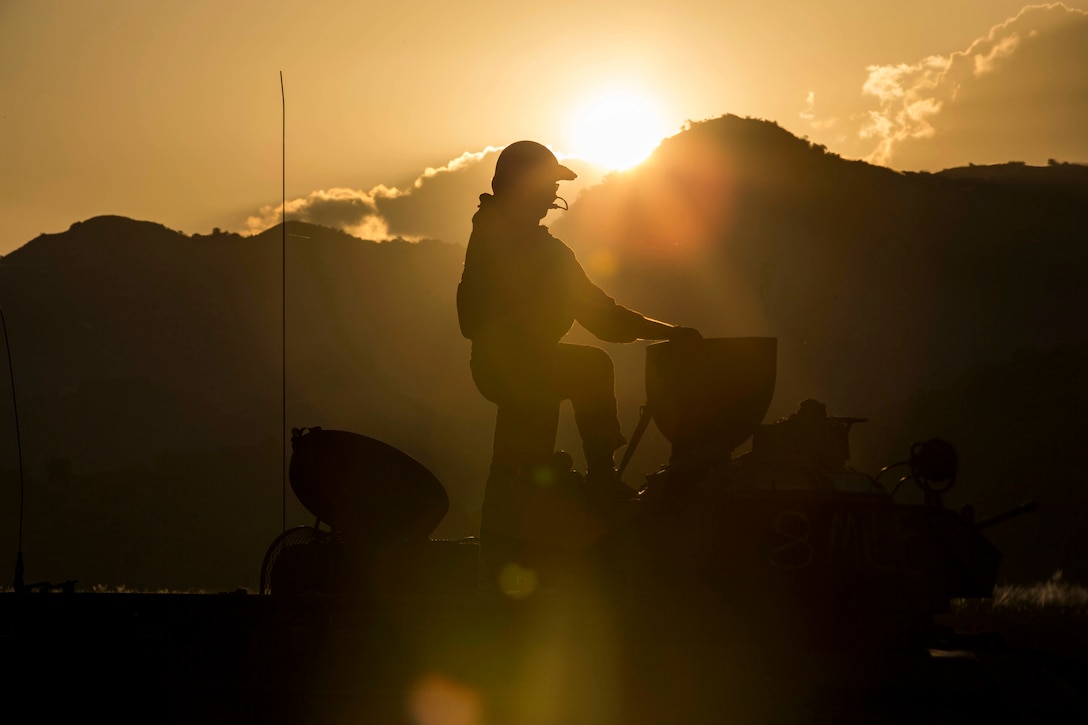 The silhouette of a Marine standing on a equipment with mountains in the background.