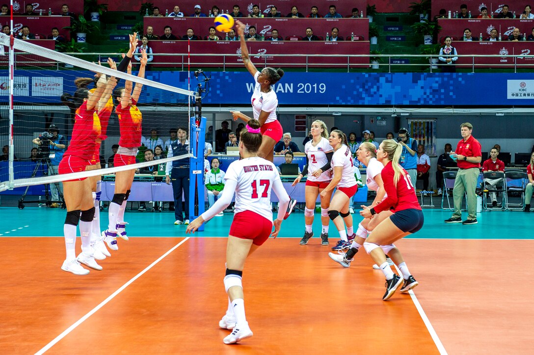 A U.S. athlete leaps and hits a ball as her teammates look on and opponents raise their arms on the other side of the net.