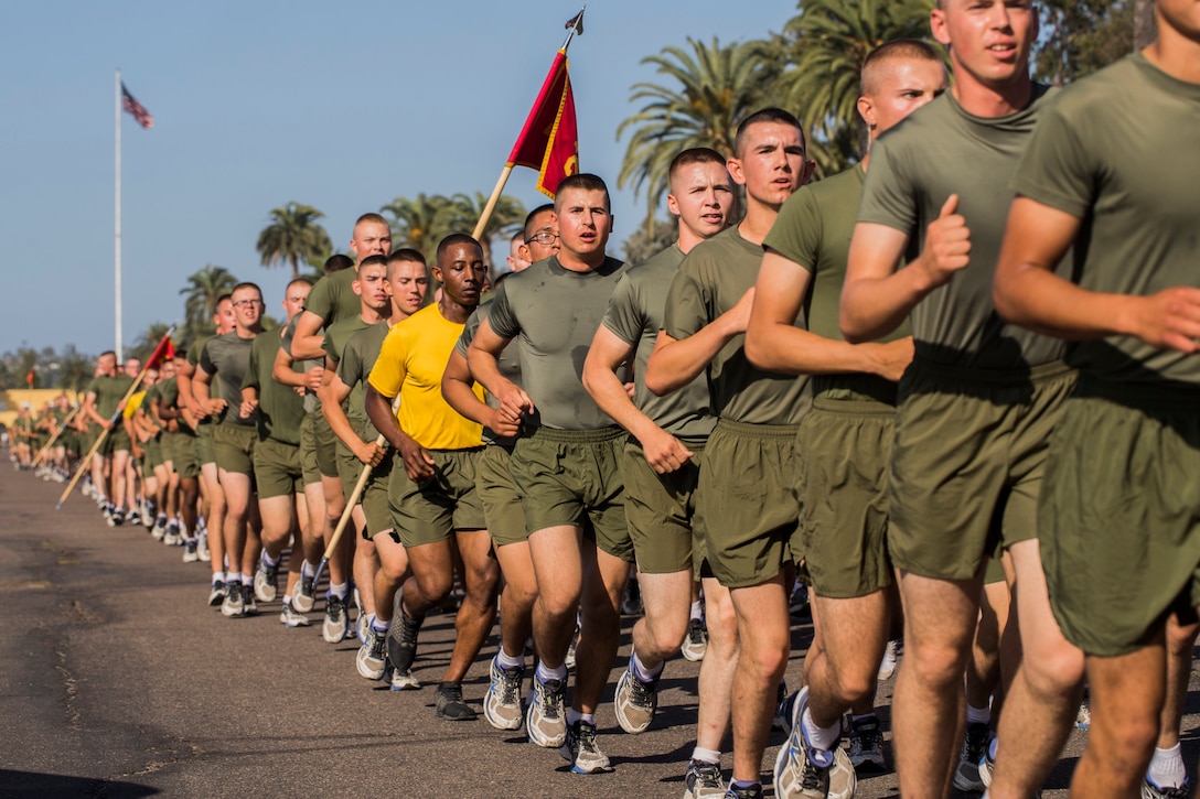 Marines jog on a street in a long line, one holding a guidon.