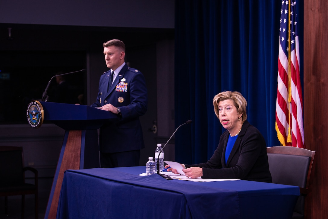 A woman sits behind a desk and speaks to reporters.