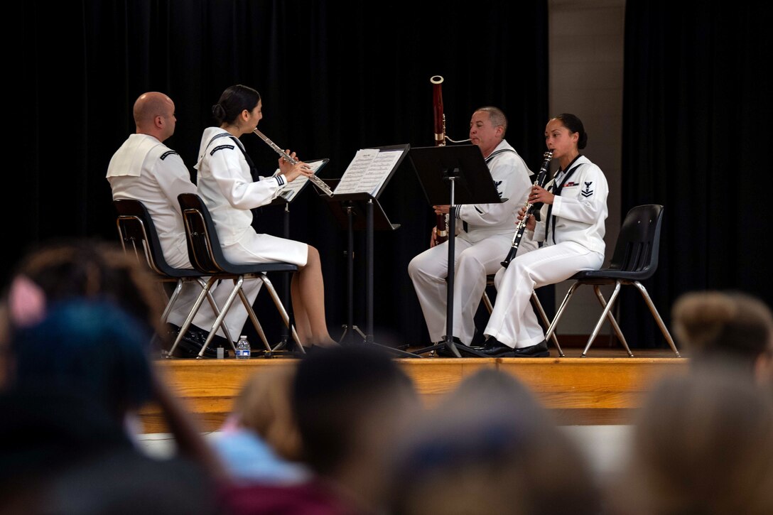 Four sailors sit on a stage playing instruments.