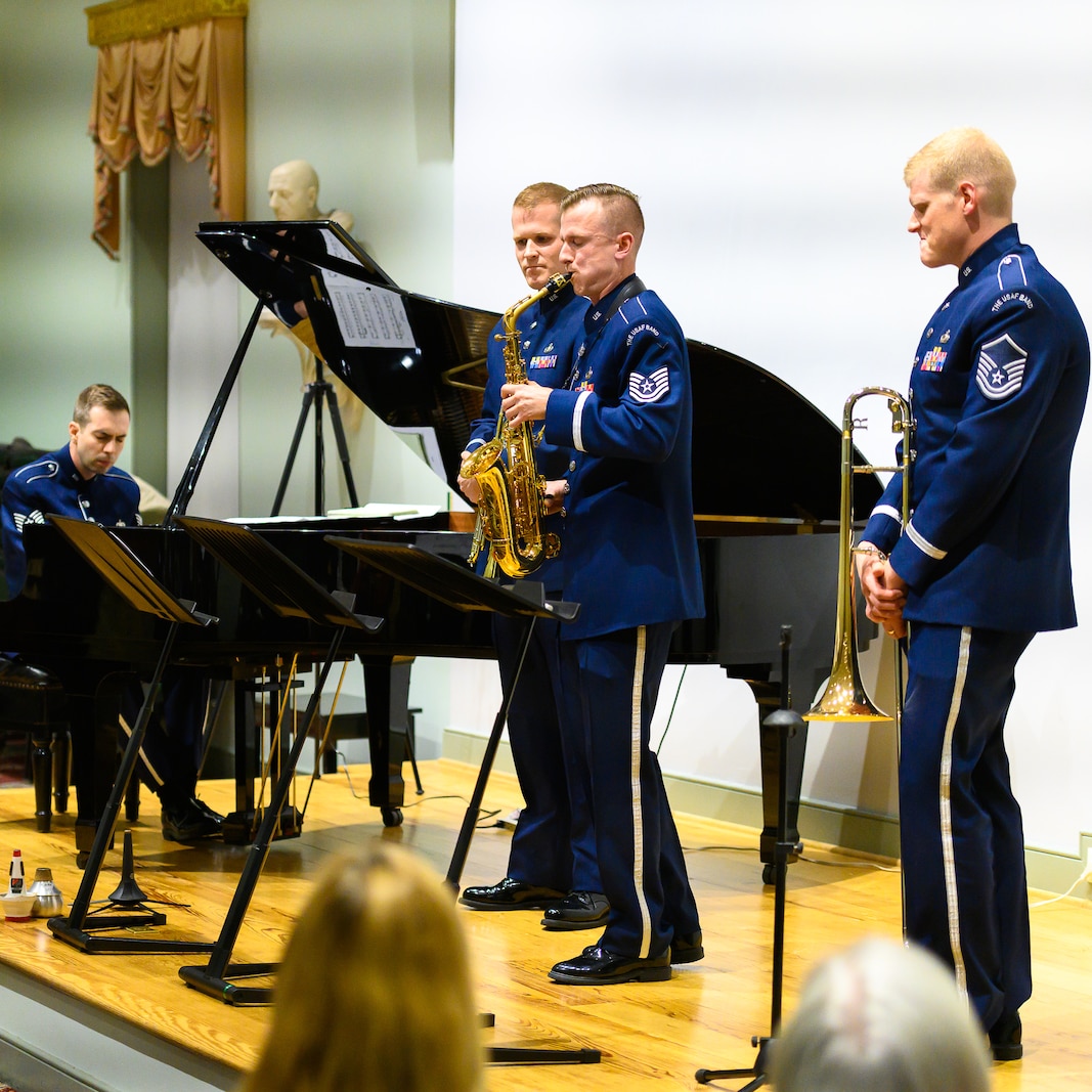 An Air Force alto saxophonist is seen flanked by two other members of the music ensemble as he performs a solo with a pianist who is seen in the far left of the picture. Each are wearing dark blue Air Force uniforms, and they are standing on a light colored wood stage.